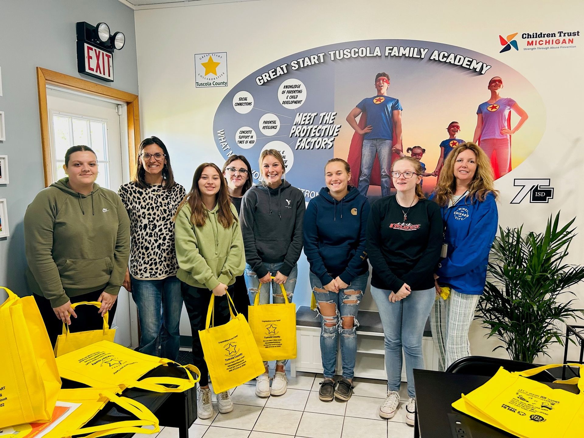 A group of people wearing casual clothing stands in front of a mural, holding yellow bags, inside a learning center.