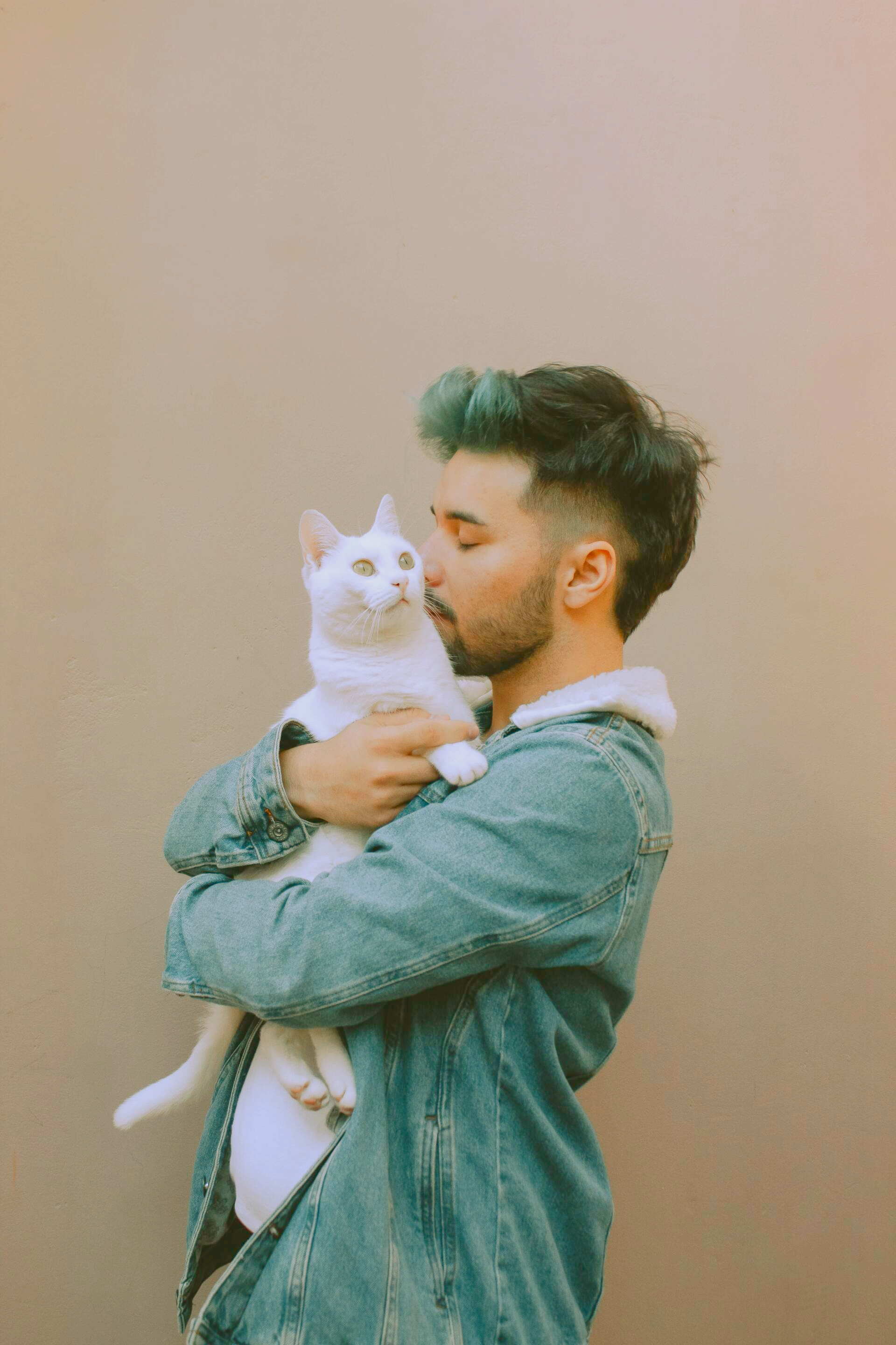 A person with teal-tinted hair wearing a denim jacket holds and nuzzles a white cat against a neutral background.