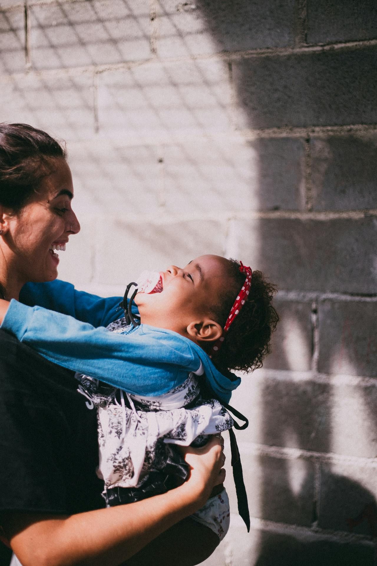 A person in a black shirt holds a laughing child wearing a blue jacket and red headband against a textured grey wall.