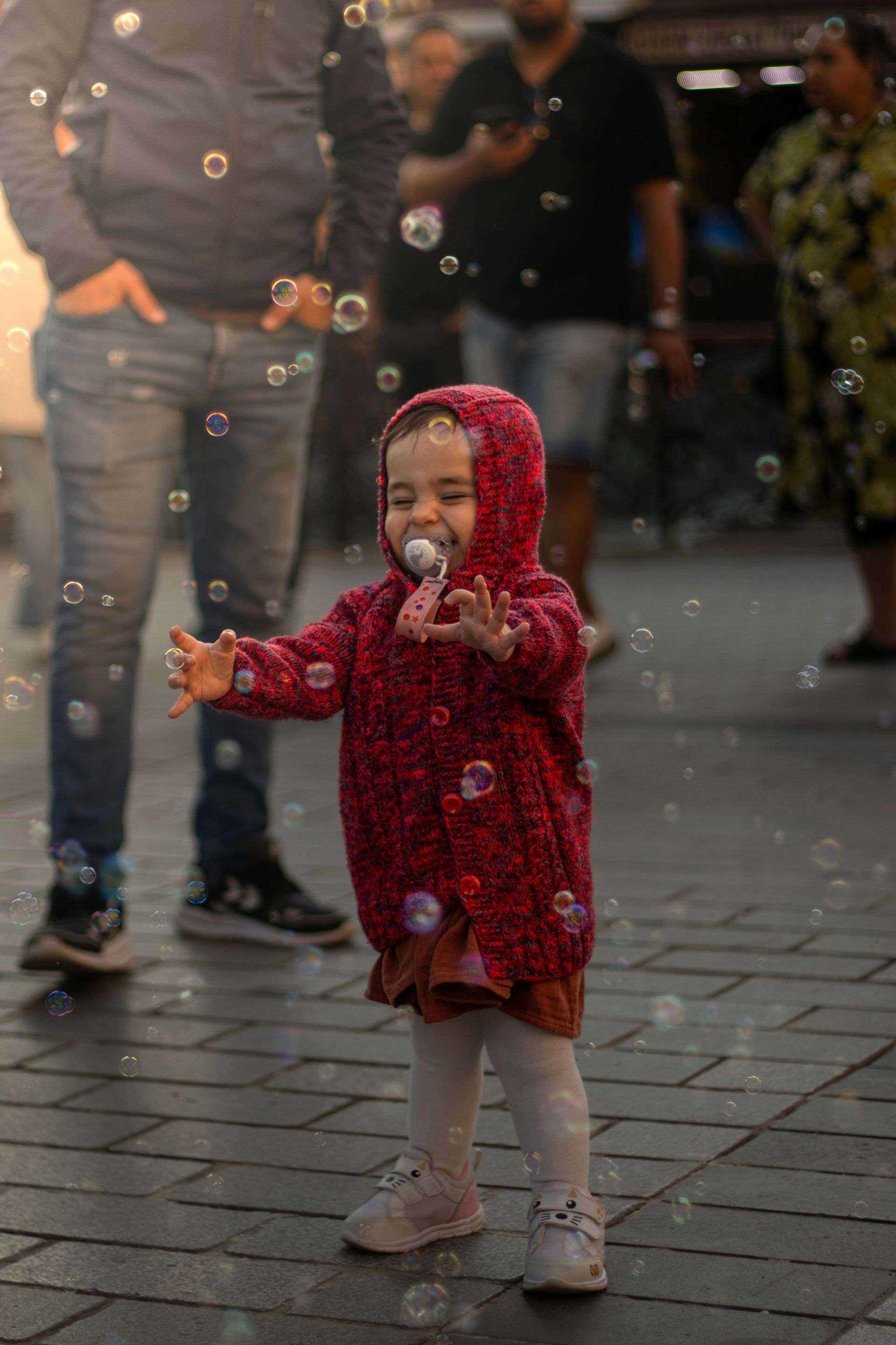 A child in a red hooded jacket reaches out toward floating bubbles on a paved city street with people standing nearby.