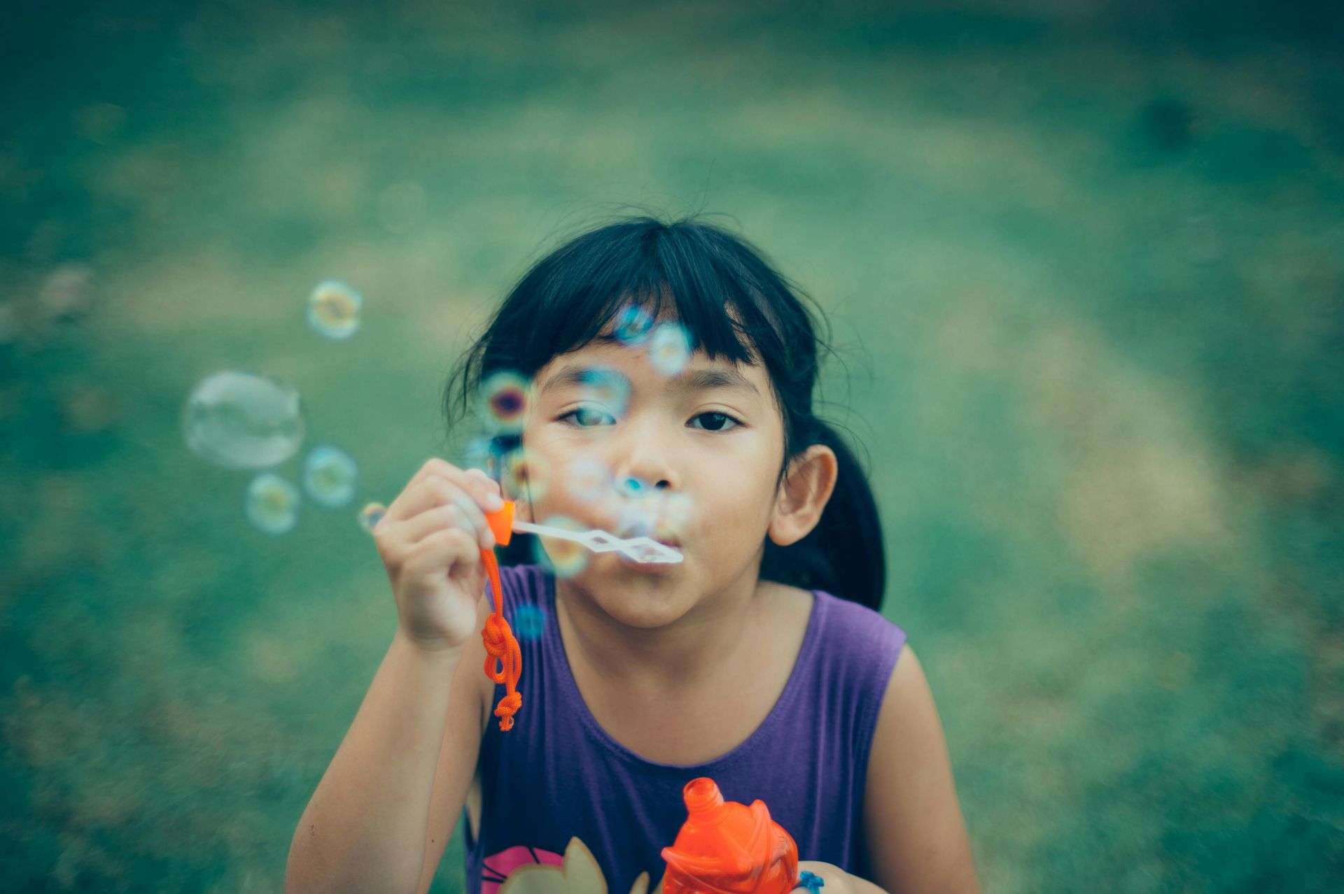 A child in a purple shirt blows bubbles from a small orange wand against a blurred green background.
