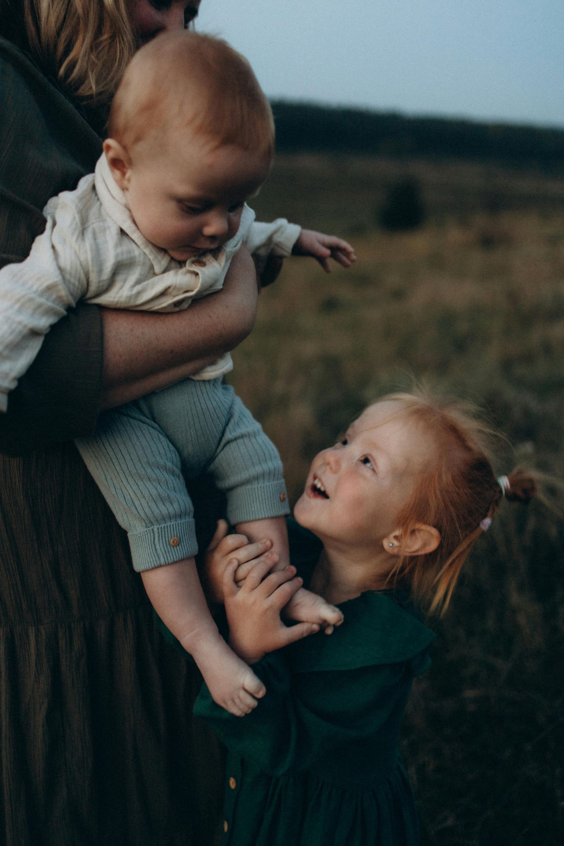 A person holds a baby while a young child looks up at the baby and touches its leg in an outdoor field at dusk.