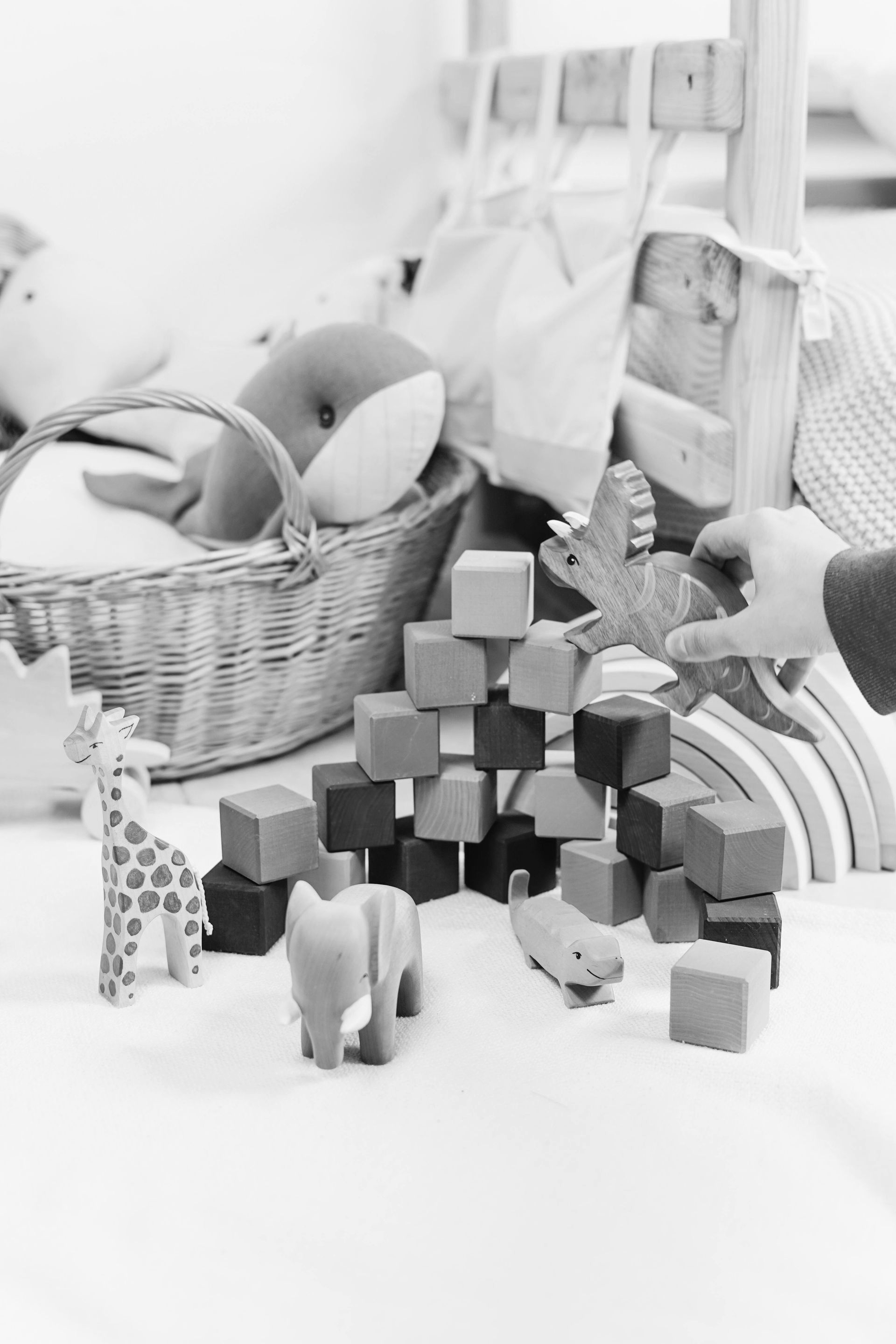 A hand places a wooden dinosaur toy on a pyramid of building blocks, surrounded by other animal figures and a basket.