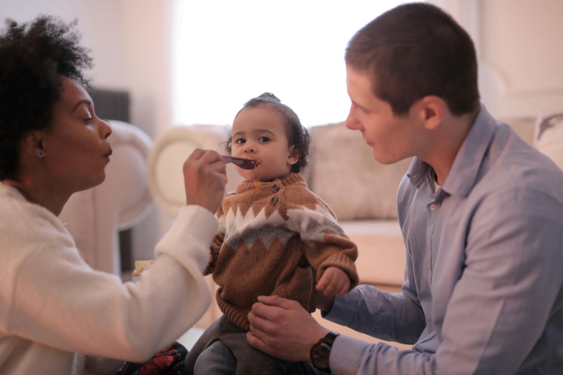 A person feeds a small child sitting between them and another adult in a softly lit living room.