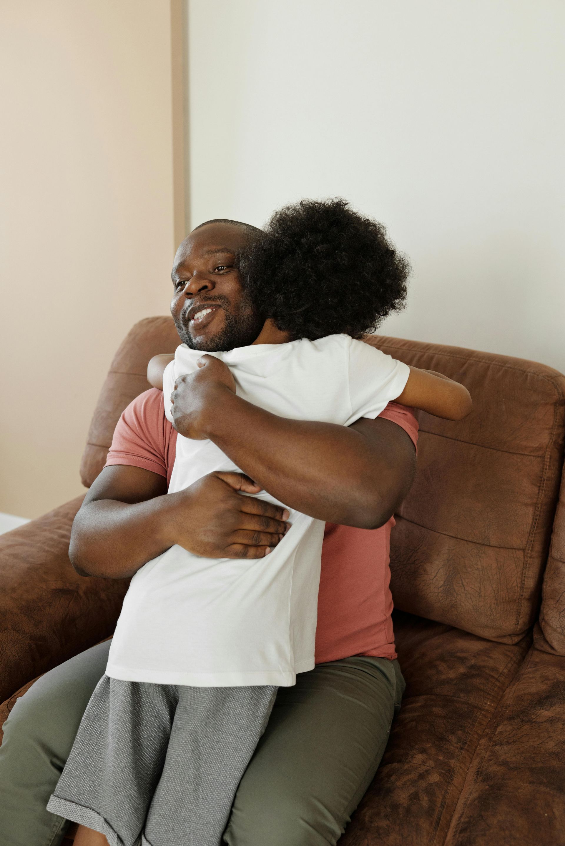 A person sitting on a brown couch hugs another individual, both smiling warmly in a bright indoor setting.