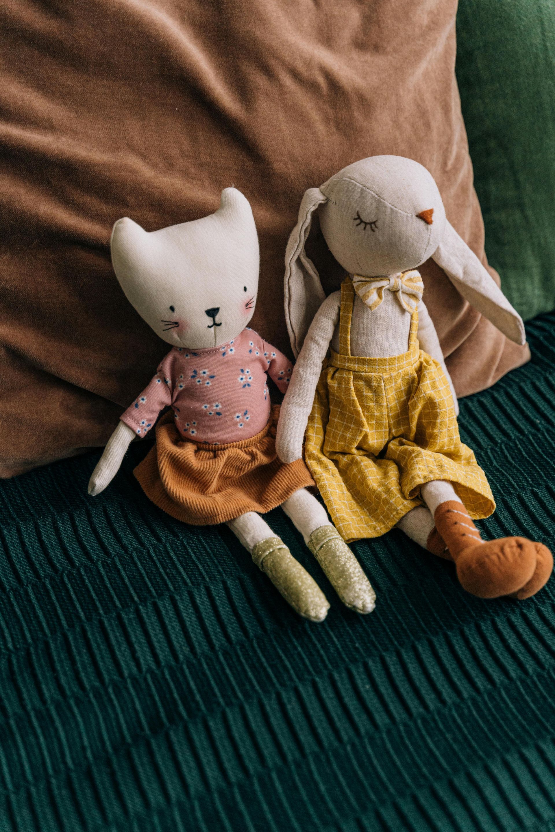 Two fabric animal dolls sit on a dark green blanket in front of a tan pillow. One is a cat, one a rabbit in yellow.