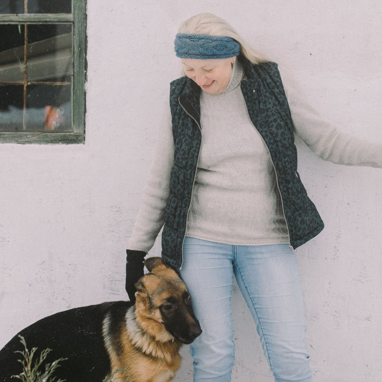 A person wearing a blue headband and a dark vest pets a German Shepherd dog against a white wall.