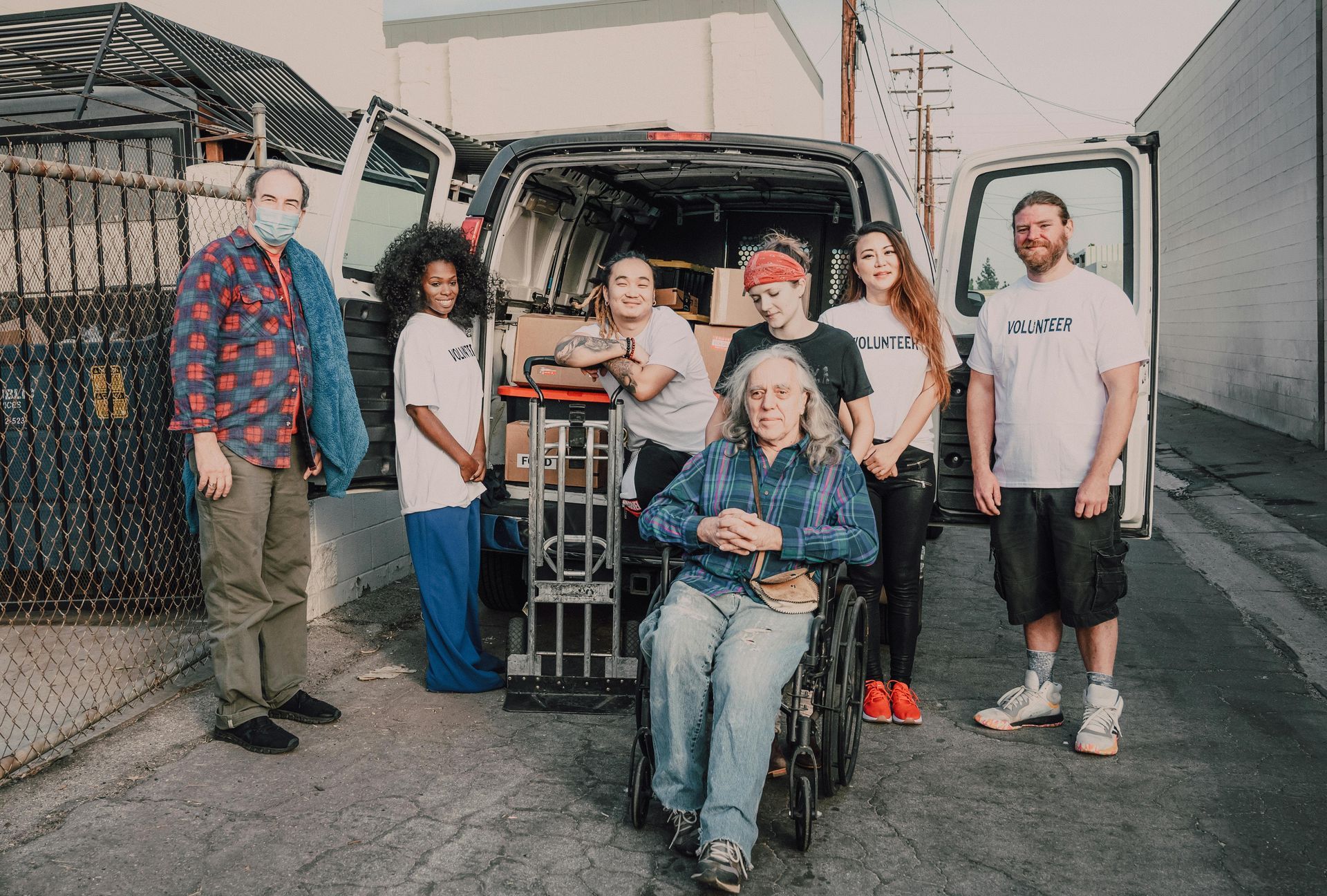 A group of people standing around a van filled with boxes, with one person in a wheelchair in front.