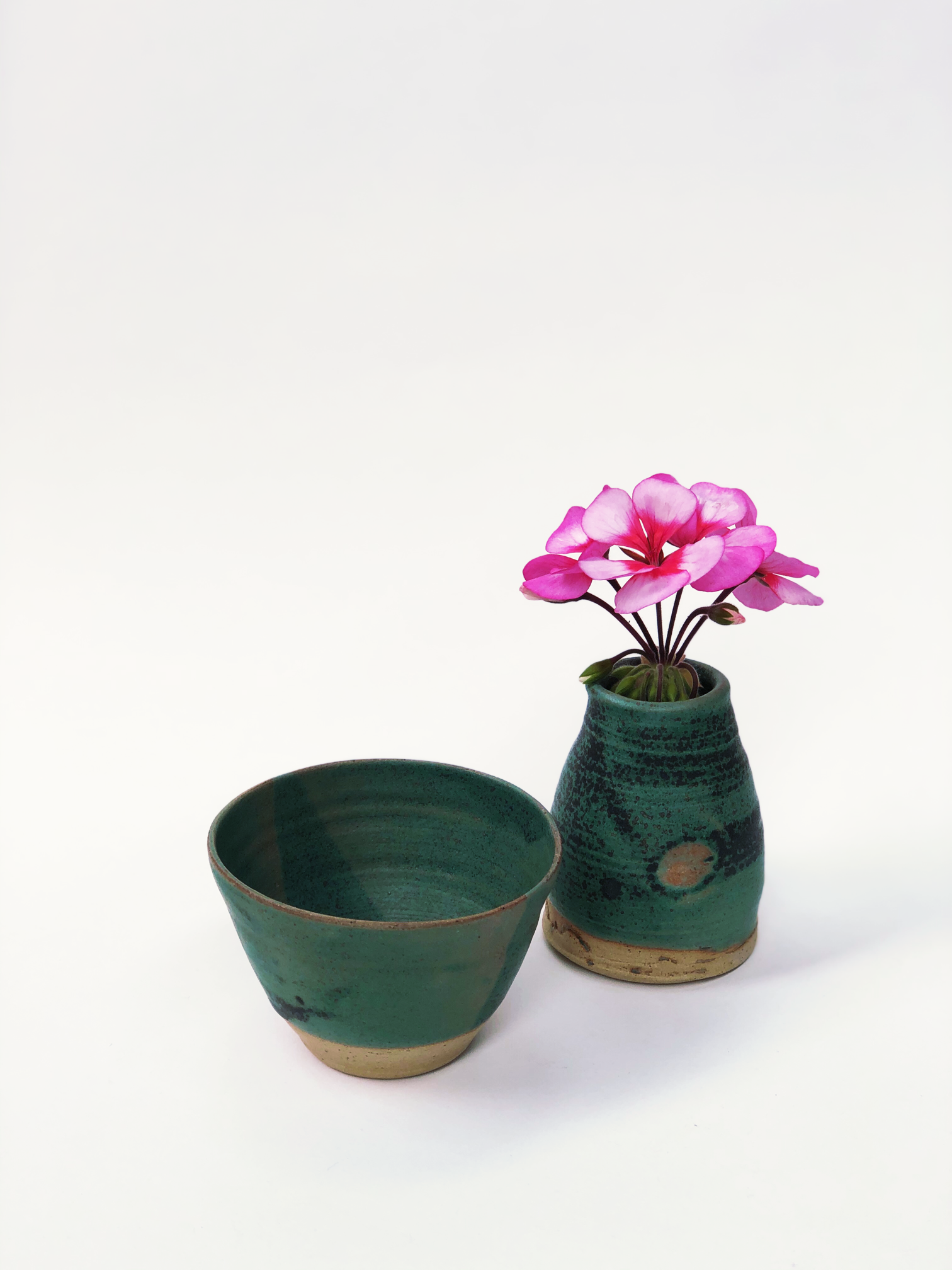 A green vase with pink flowers in it next to a green bowl.