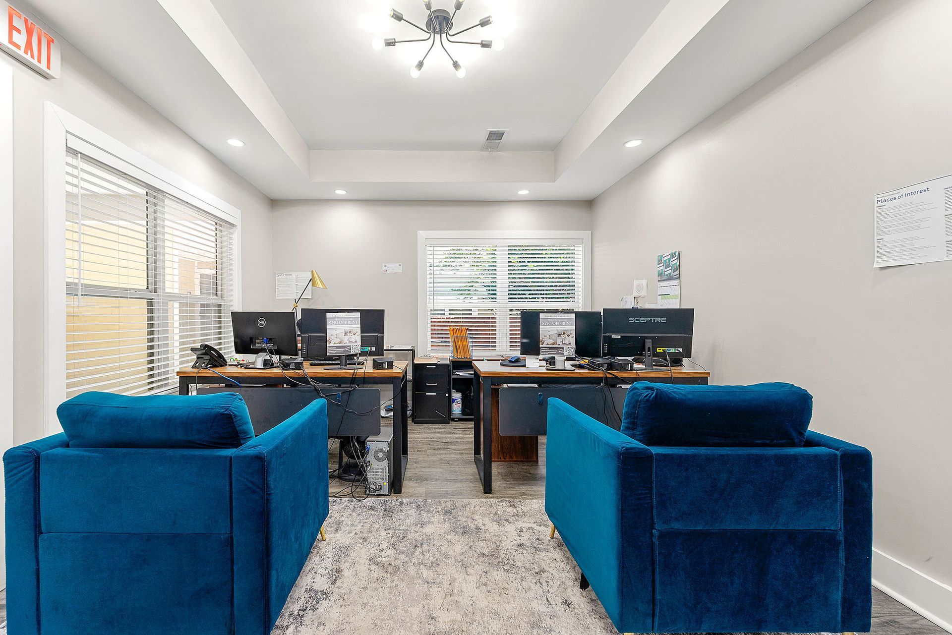 Modern office lounge with blue chairs, desks, computer monitors, and large windows under bright ceiling lights