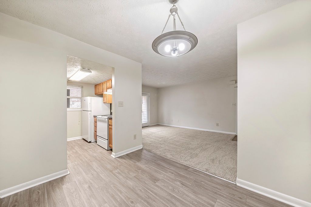 Empty apartment interior with light wood floors, carpeted living area, and view into kitchen.