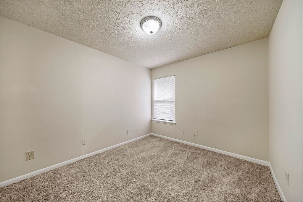 Empty beige bedroom with carpet, white walls, a ceiling light, and a small window.
