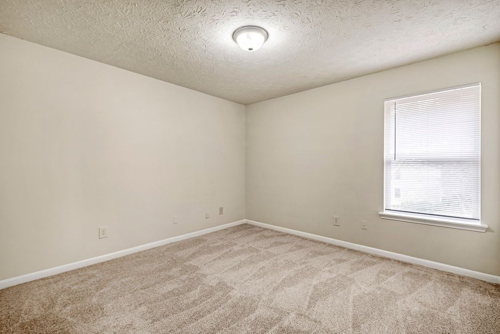 Empty beige bedroom with carpet, ceiling light, and one window with a blind.