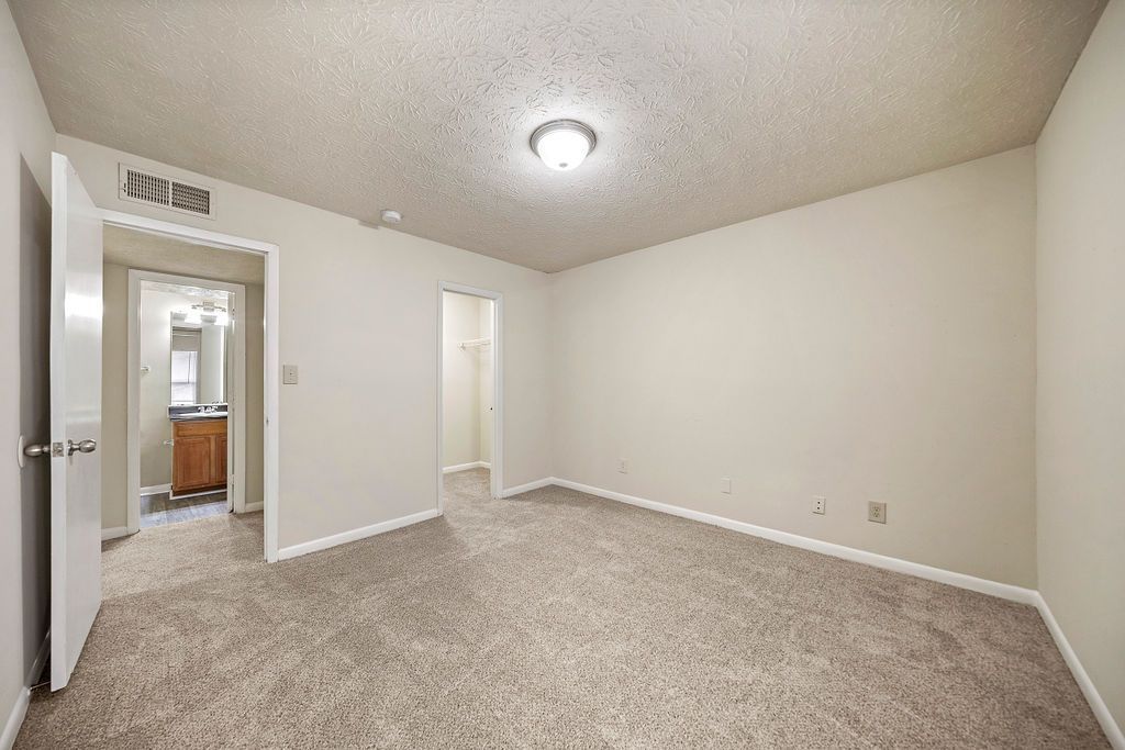 Empty beige carpeted bedroom with white walls, ceiling light, and open doorway to a bathroom