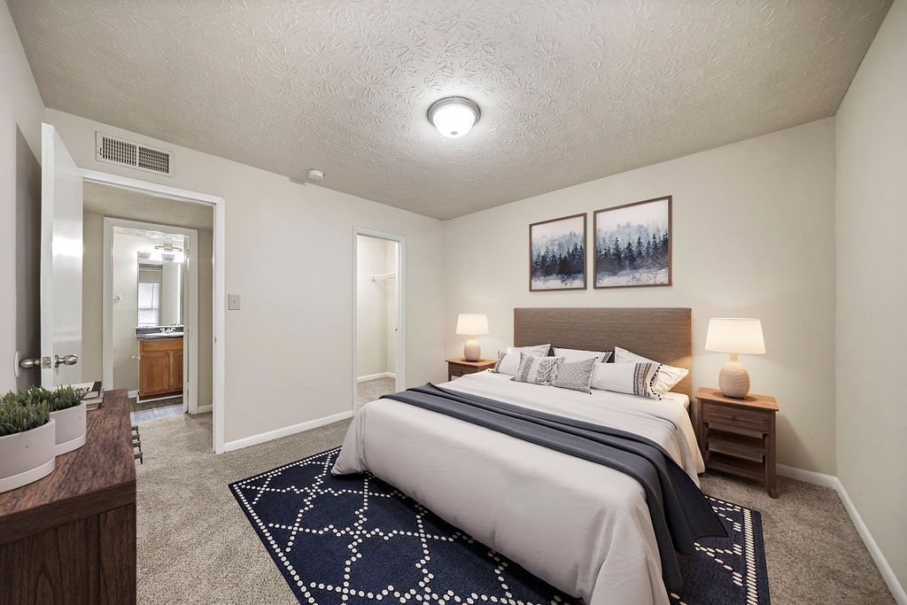 Spacious neutral bedroom with a king bed, dark blue rug, nightstands, wall art, and an open doorway.
