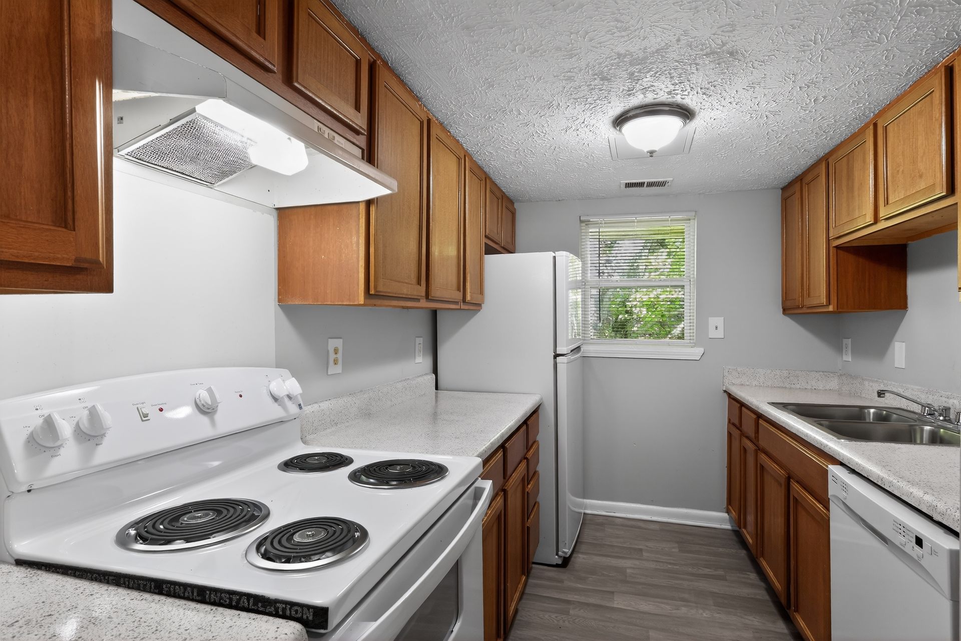 Small galley kitchen with white appliances, wood cabinets, and a window at the end.