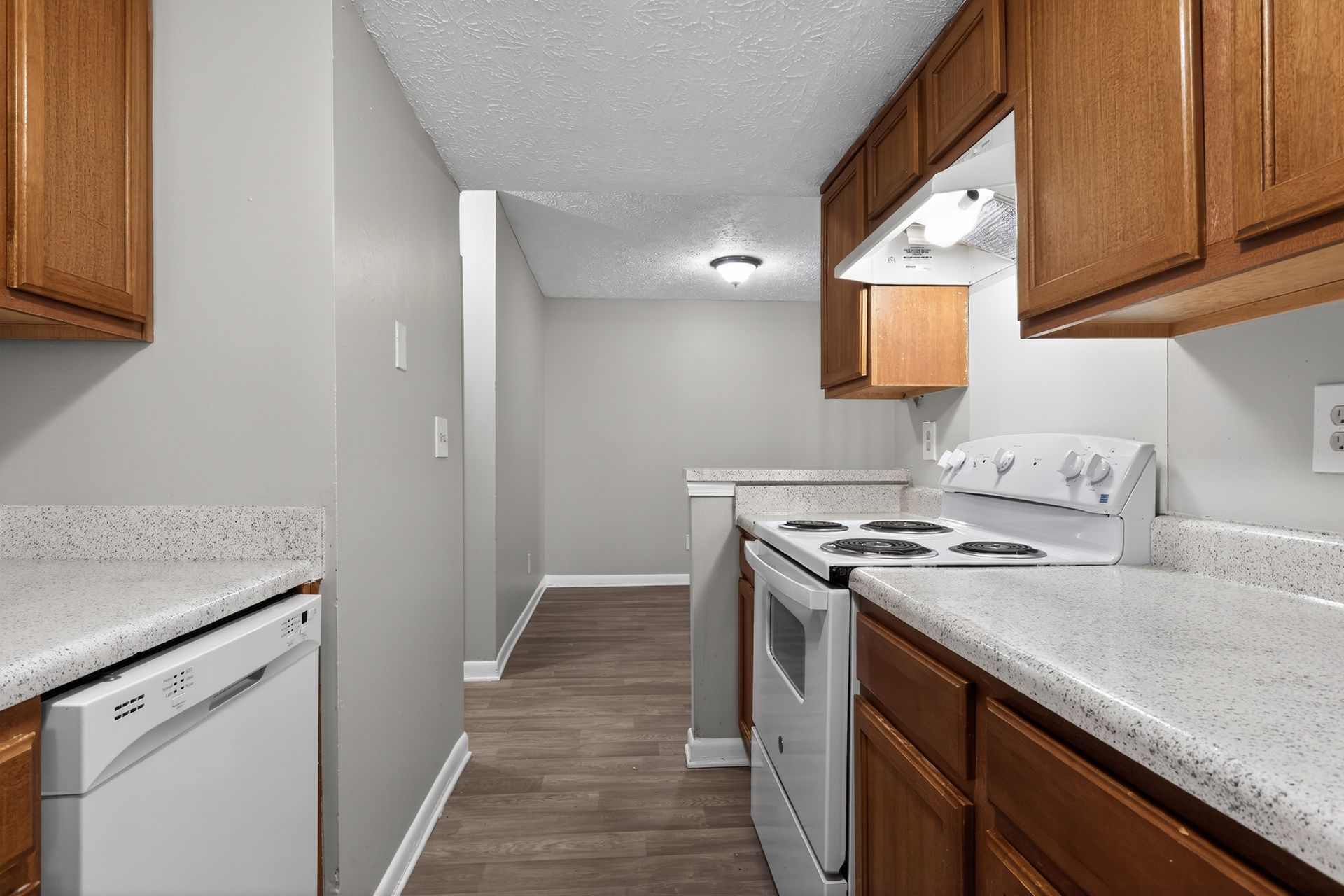 Empty kitchen with white appliances, speckled counters, and wood cabinets in a narrow hallway layout