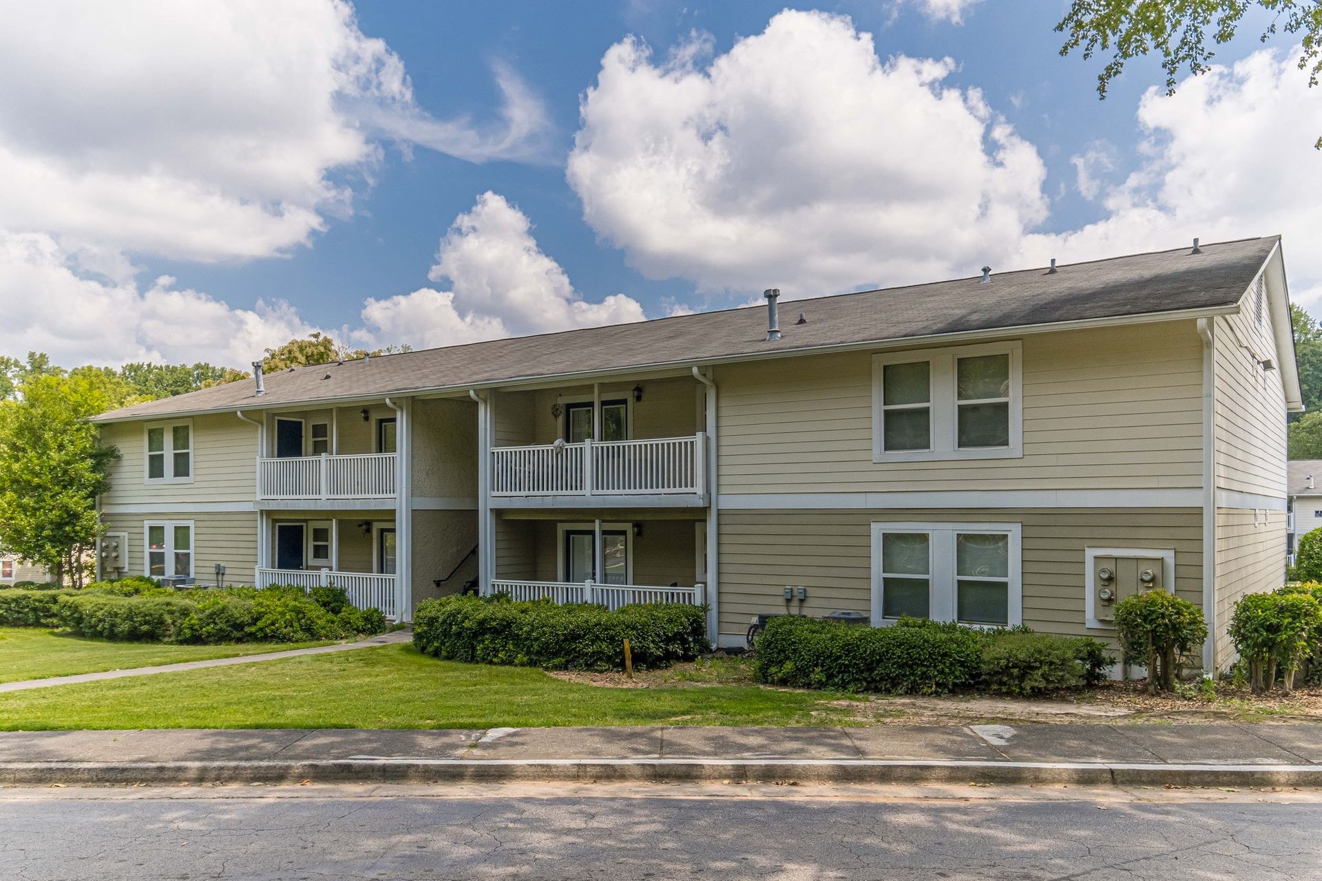 Two-story beige apartment building with balconies, white trim, and green landscaping under a partly cloudy sky