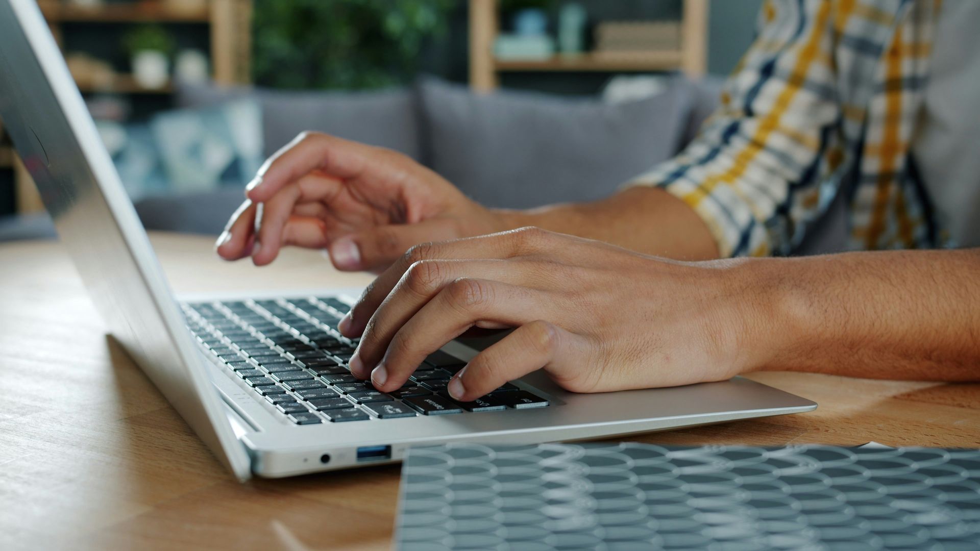 Hands typing on a silver laptop at a wooden table in a bright indoor setting.