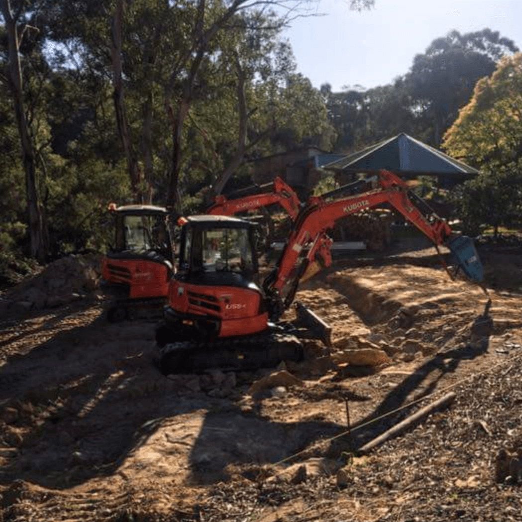 Two orange excavators working on a dirt lot near trees and a house
