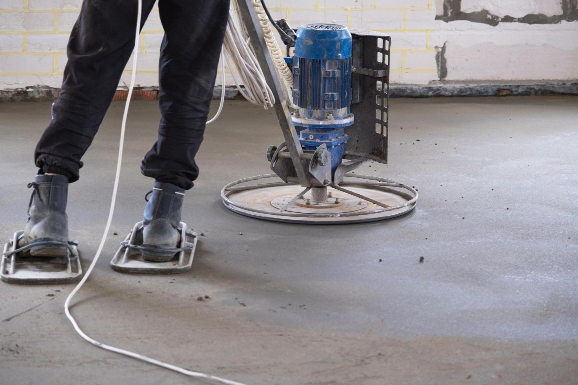 Worker using a floor grinder on a concrete surface indoors, with a power cord trailing nearby