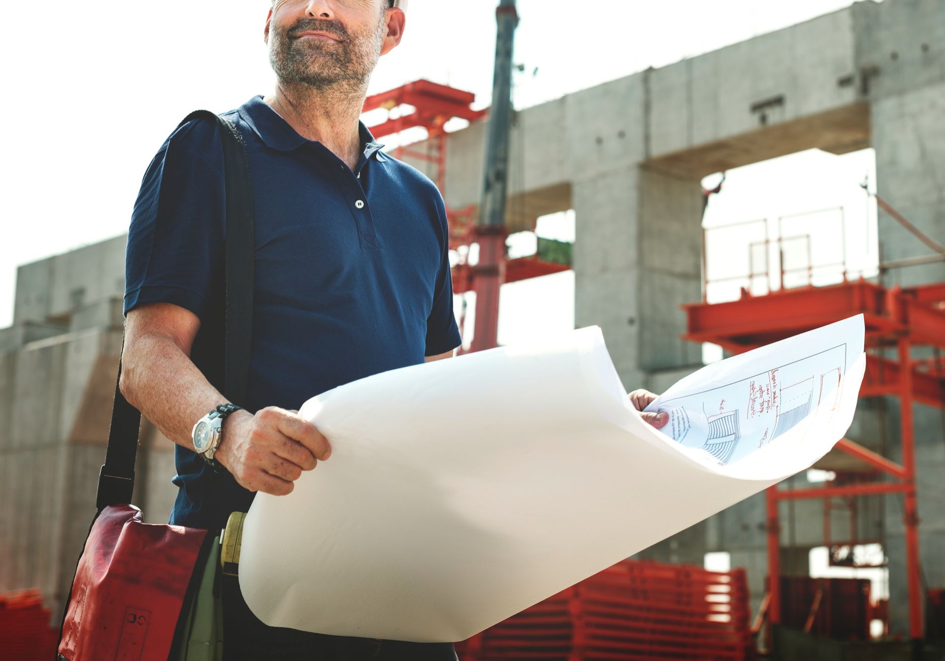 Man in navy shirt holding rolled blueprints at a construction site with red scaffolding and concrete walls