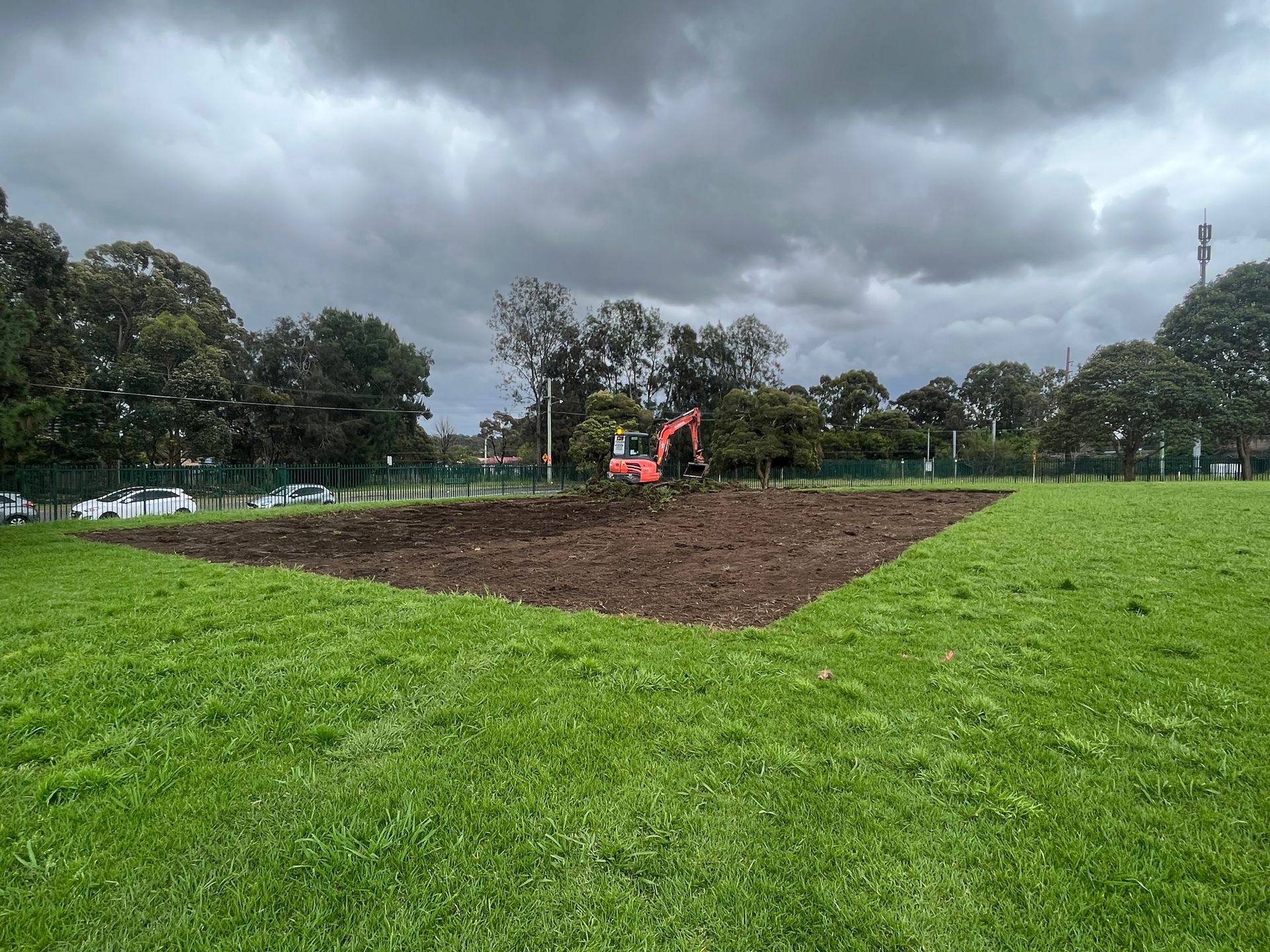 Tractor tilling a brown field beside a green park under a cloudy sky