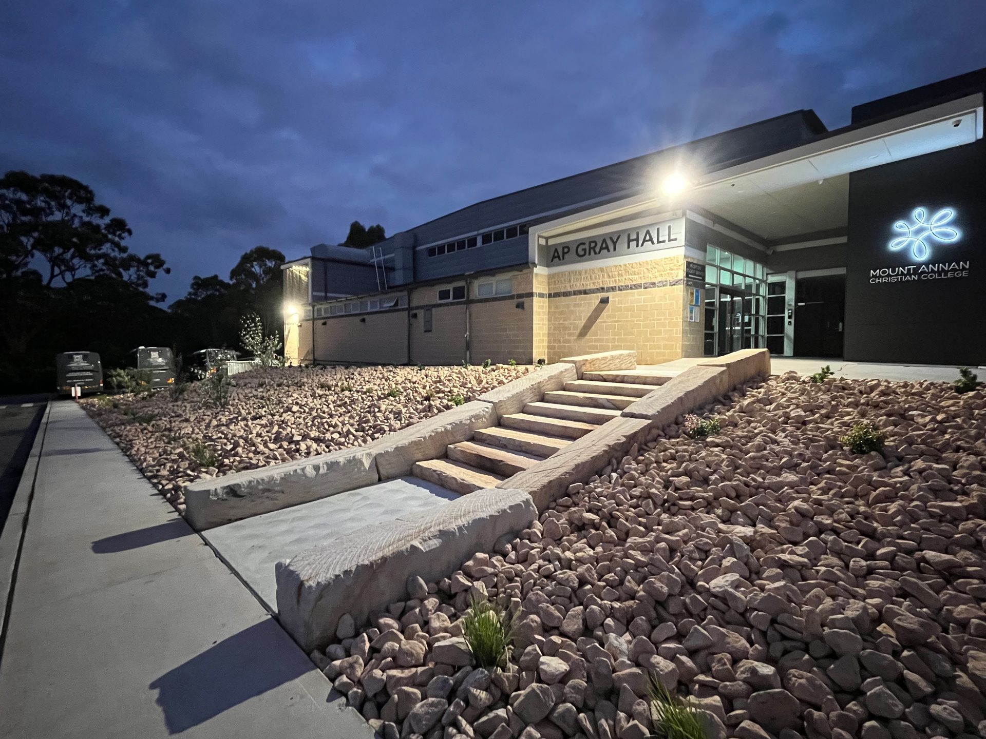 Modern building entrance at dusk with lit stairs, glass doors, and landscaped rock-covered slopes