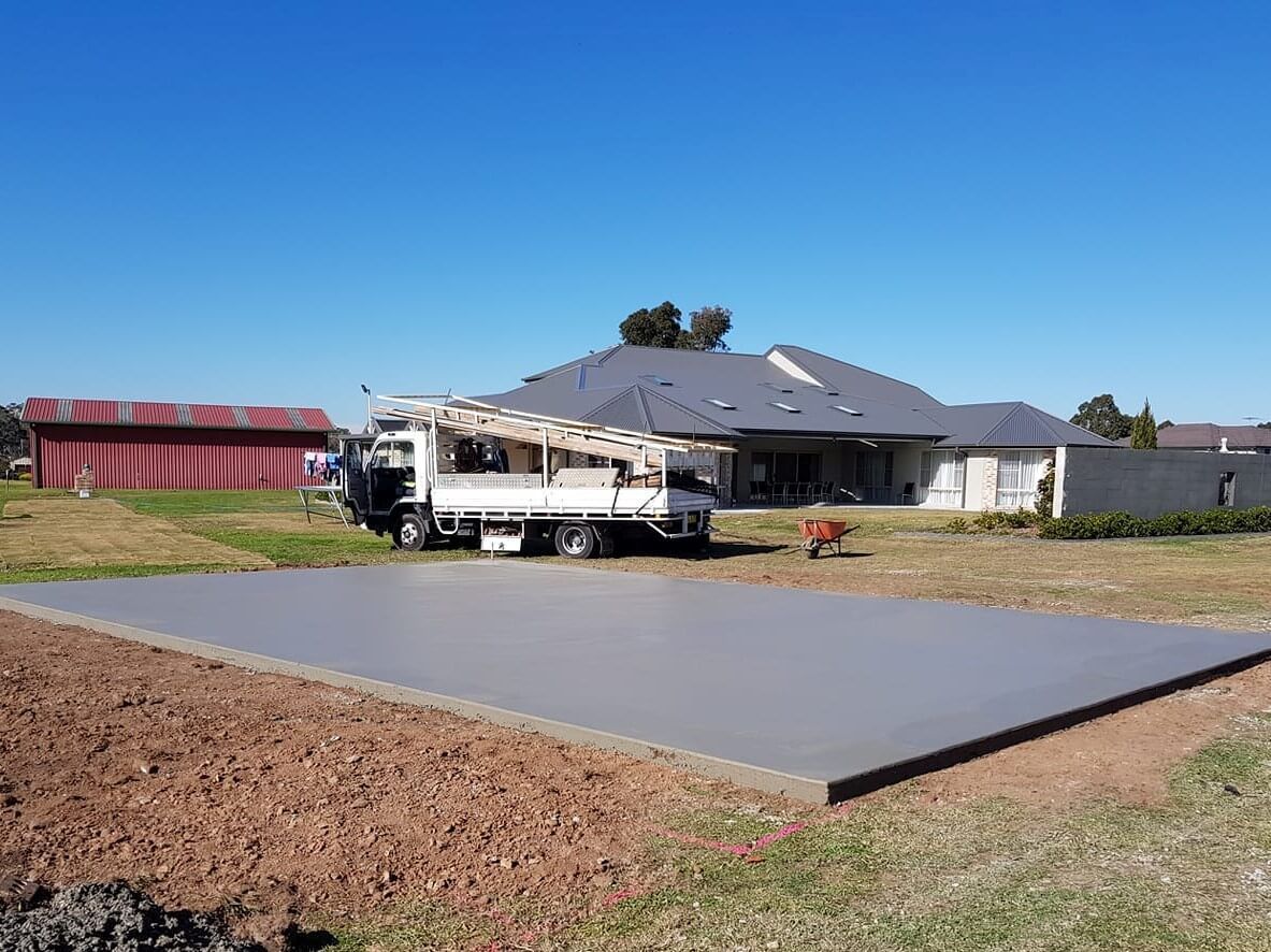 Concrete slab being poured at a suburban house, with a truck and workers nearby under a blue sky