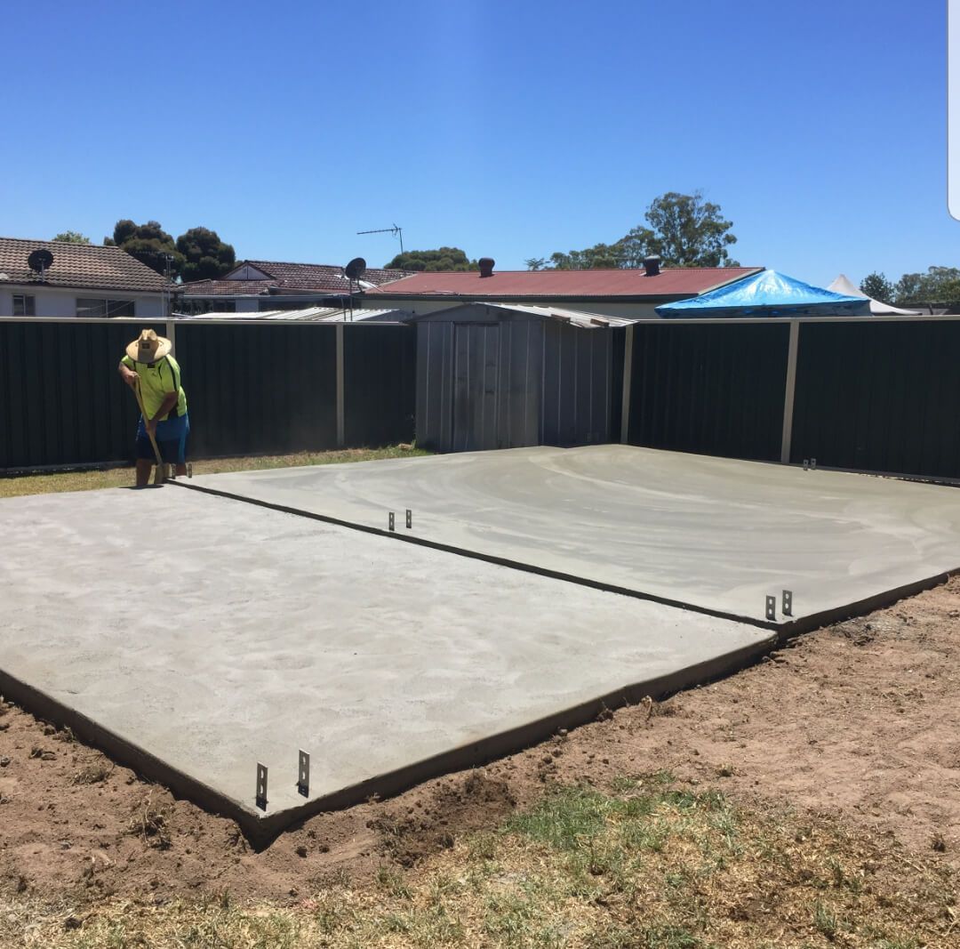 Fresh concrete slab foundation in a fenced backyard, with a worker on the left under a clear blue sky