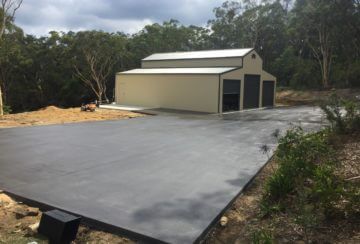 Concrete driveway leading to a beige metal garage with two dark doors, surrounded by trees and dirt