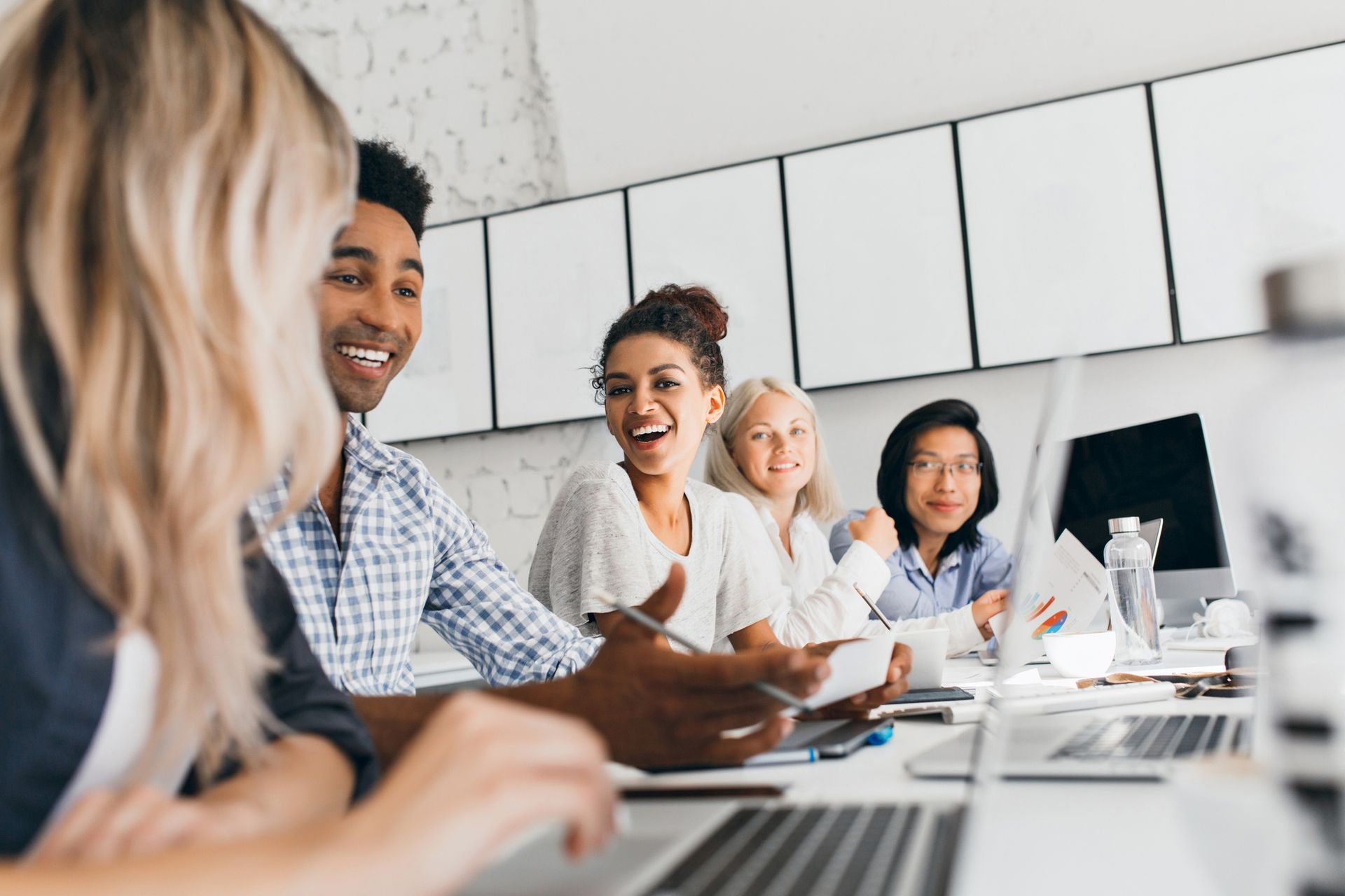 People in a bright meeting room working and smiling around a conference table with laptops and papers