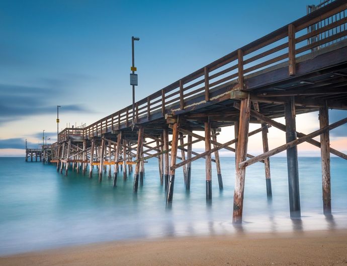 Wooden pier extending over calm blue water at dusk, with gentle waves and sandy shore in foreground