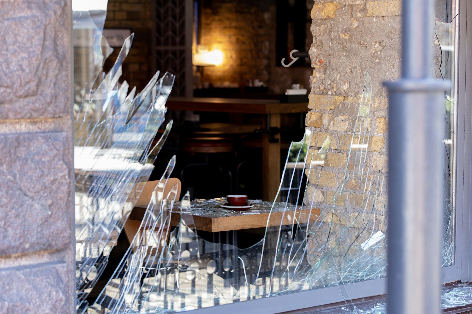 Broken glass storefront with shattered window panes and a visible table inside an empty stone building.