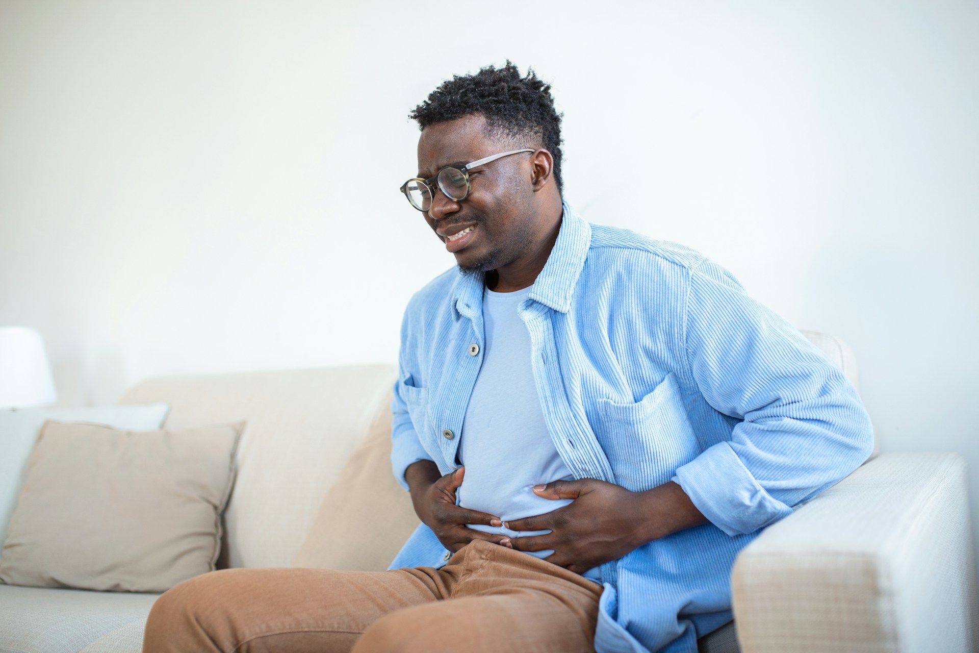 Man sitting on a couch, clutching his abdomen and grimacing in discomfort.