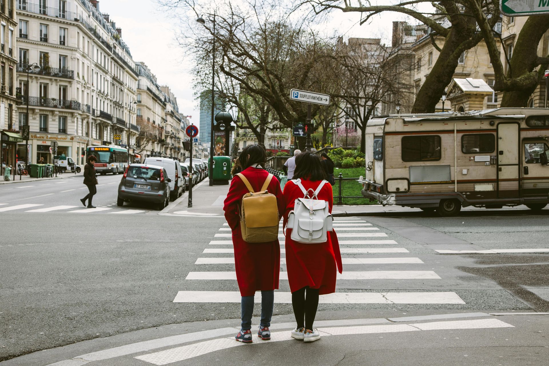 Two people in red coats cross a zebra crossing with backpacks in a city street near buildings and a parked camper van