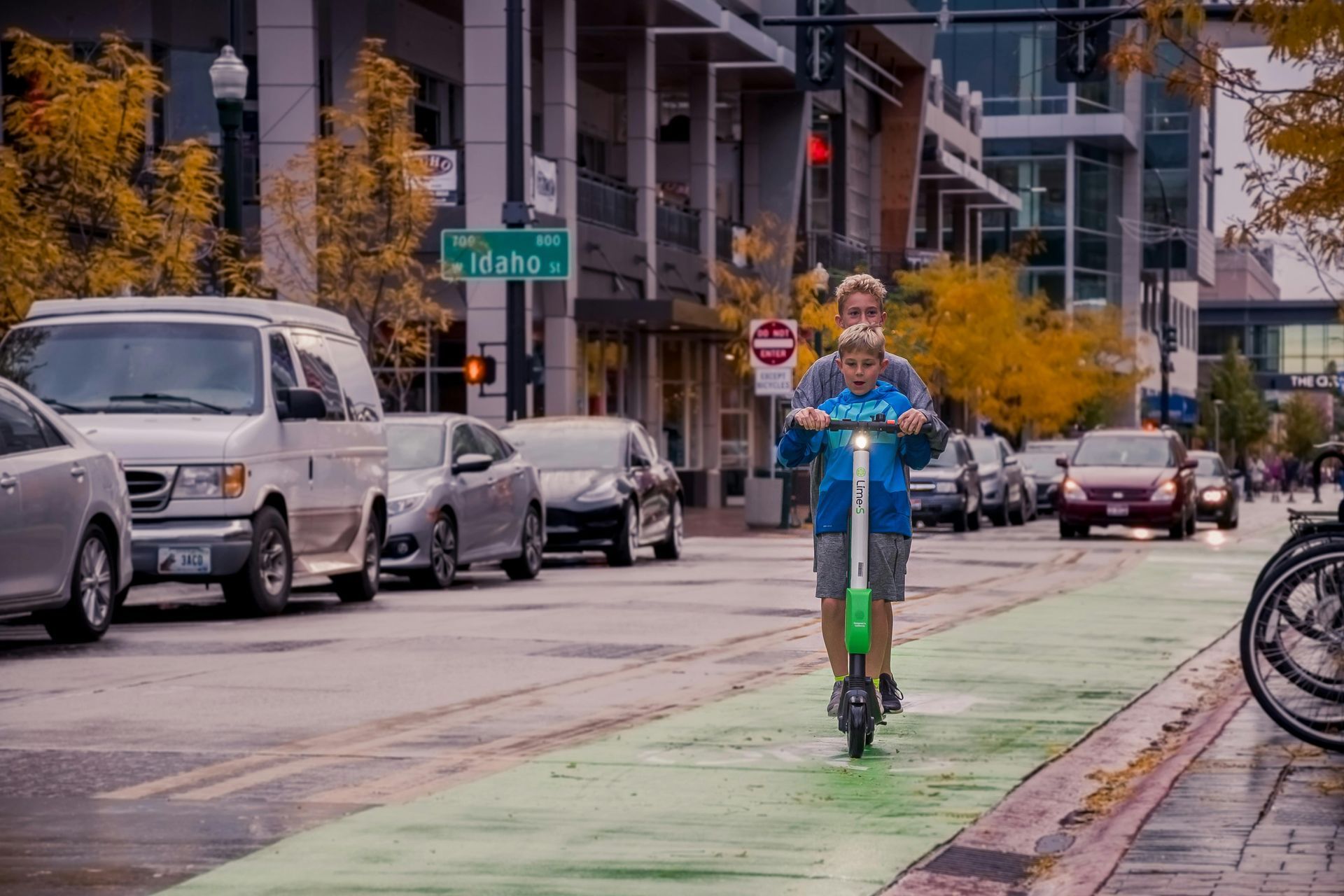 Person riding a blue scooter in a green bike lane on a city street, with parked cars and buildings nearby.