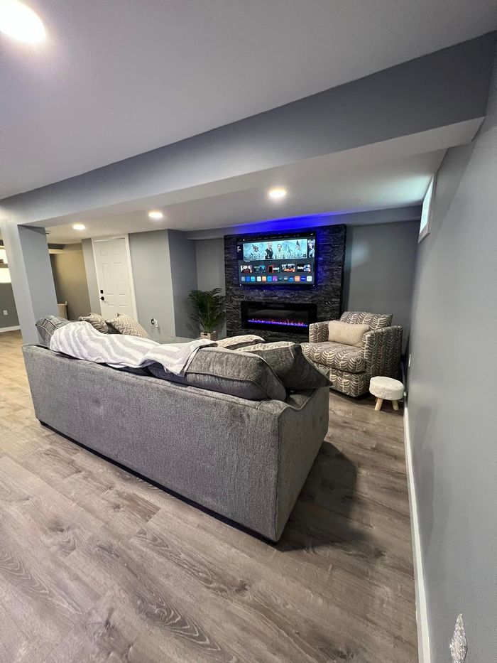 A modern living room with gray walls and flooring. A large gray sofa faces a fireplace with a lit TV.