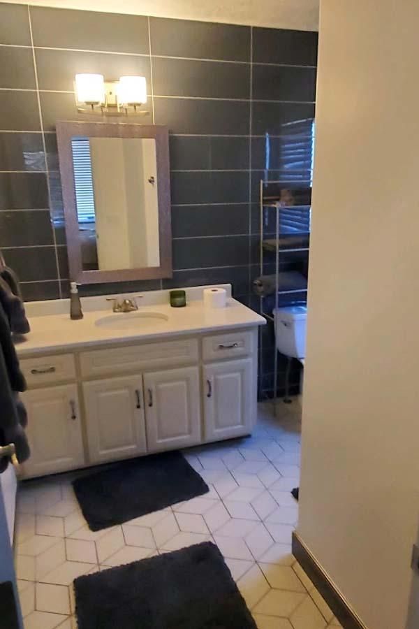 Bathroom with white vanity and sink, dark gray tiled walls, and a toilet. Black bath mats on the tiled floor.