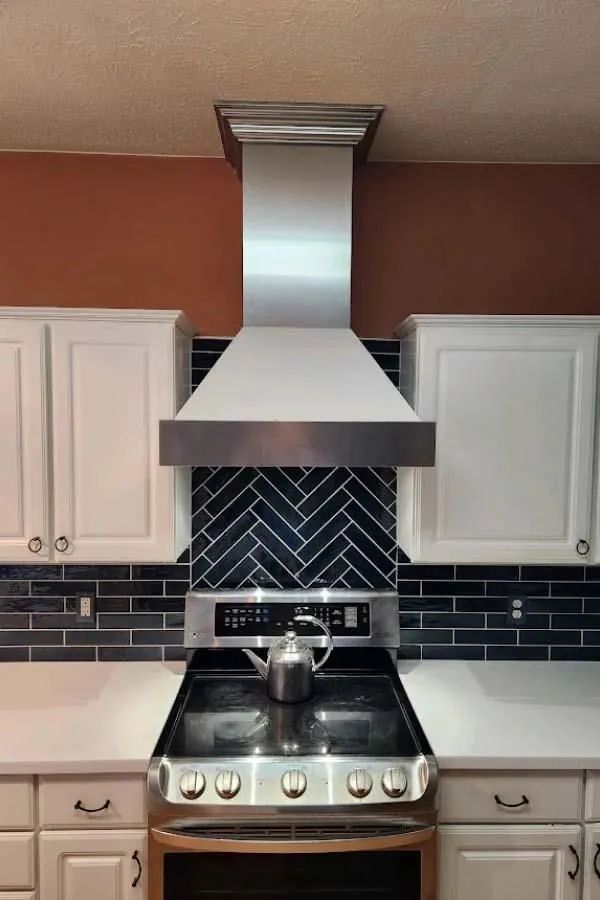 Kitchen with white cabinets, stainless steel range hood, blue herringbone backsplash, and stovetop with a kettle.