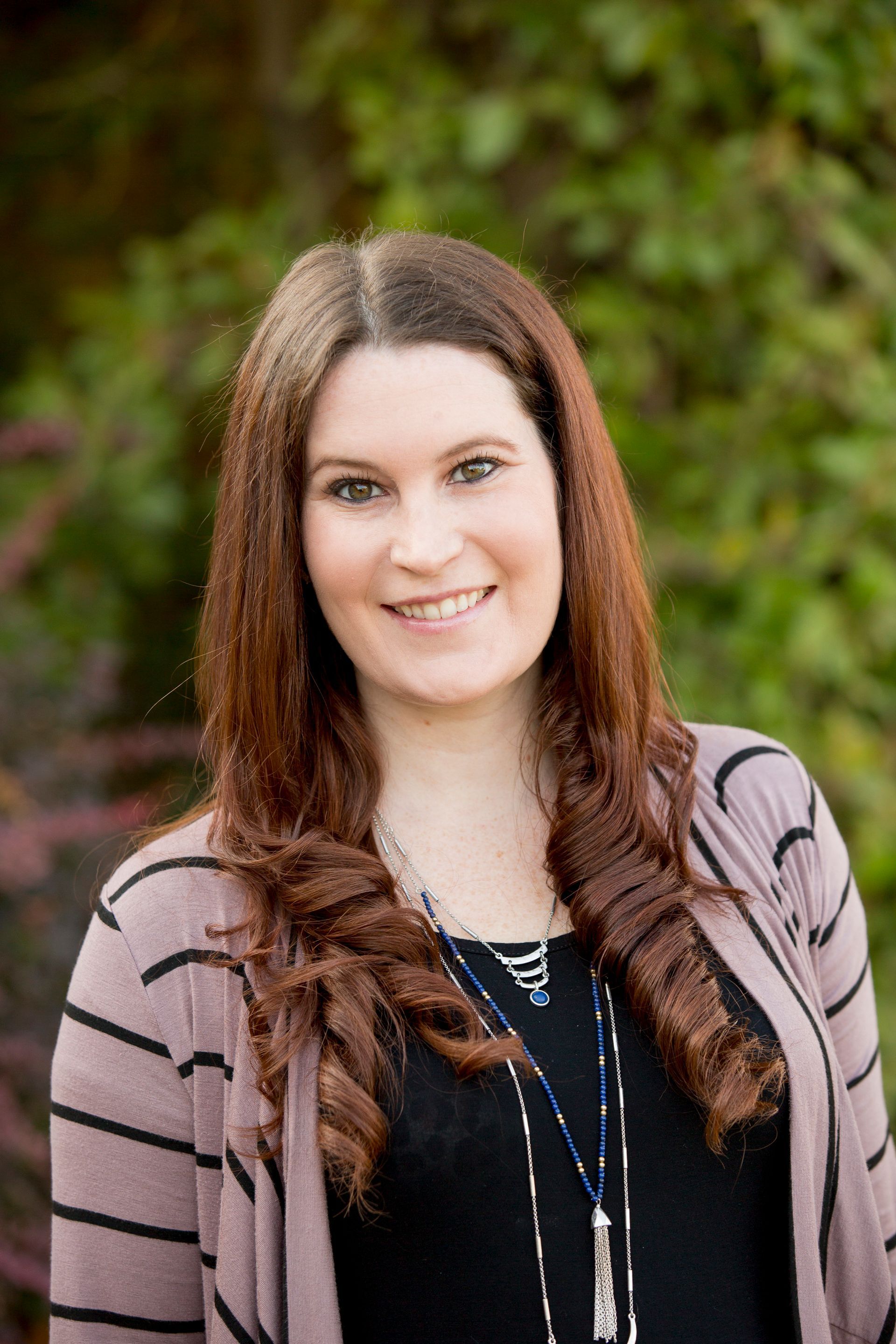 Portrait of a smiling woman with long brown hair, wearing a black top and pink cardigan outdoors.