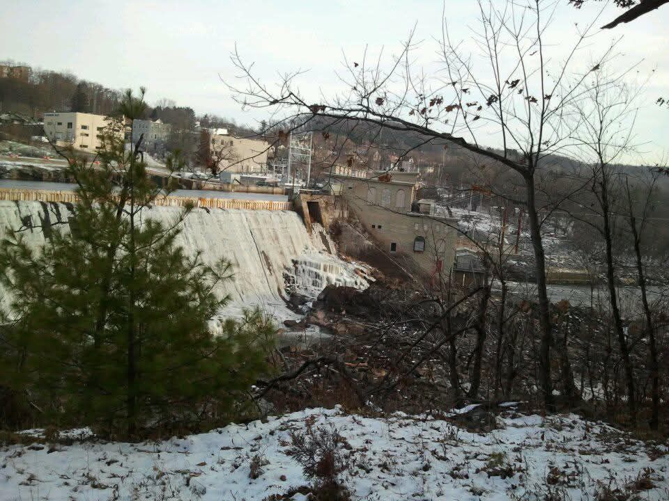 Snowy riverside view of a dam with white water, bare trees, and buildings on the hillside