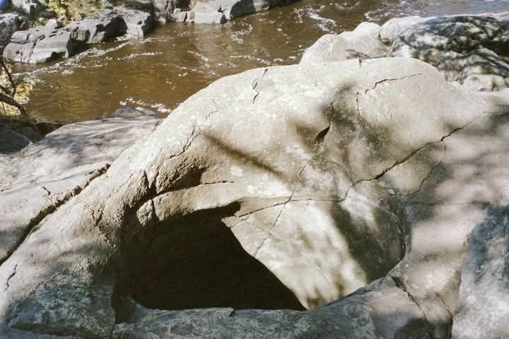 Large white rock with a dark hollow beside a muddy stream, with rocky banks in the background