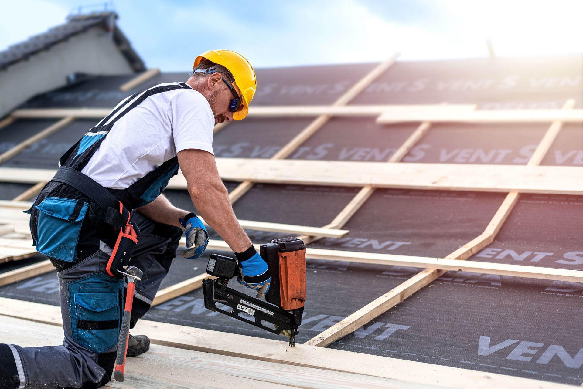 Worker in a yellow hard hat installs roofing underlayment with a nail gun.
