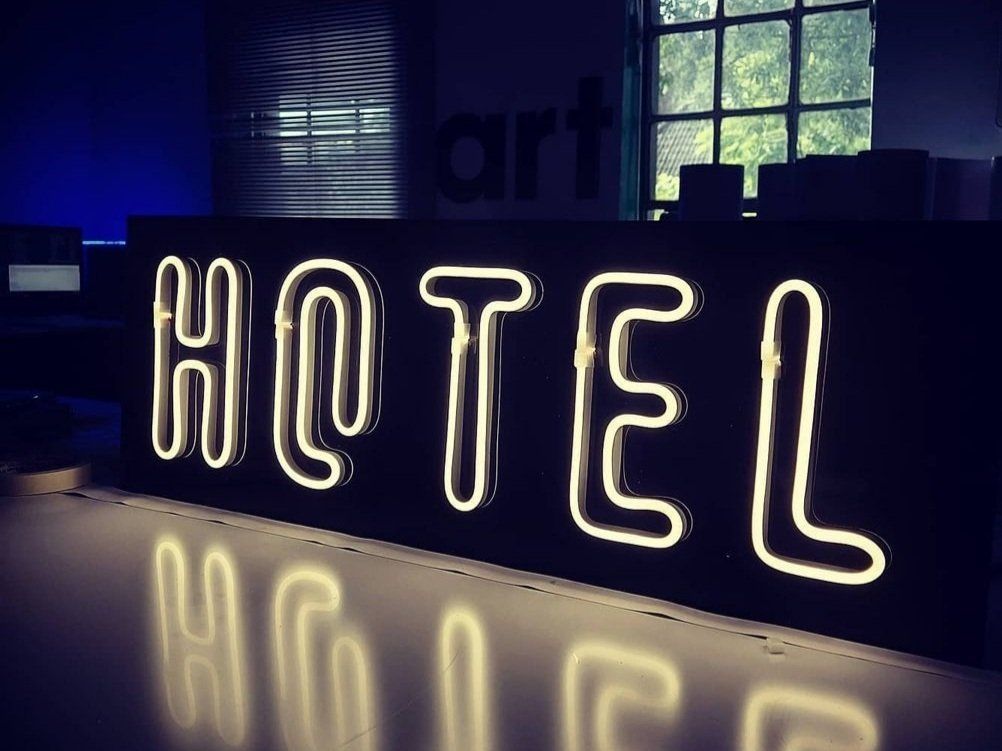 Neonschild mit der Aufschrift „HOTEL“ auf einer weißen Fläche, mit einem Fenster im Hintergrund.