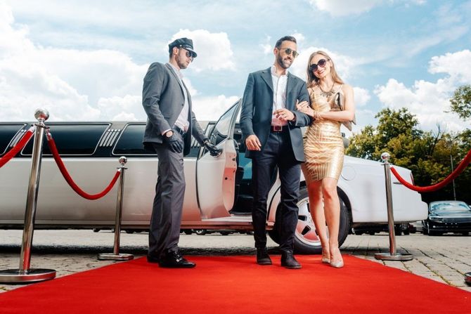 Three stylish people on a red carpet beside a limousine, posing outdoors