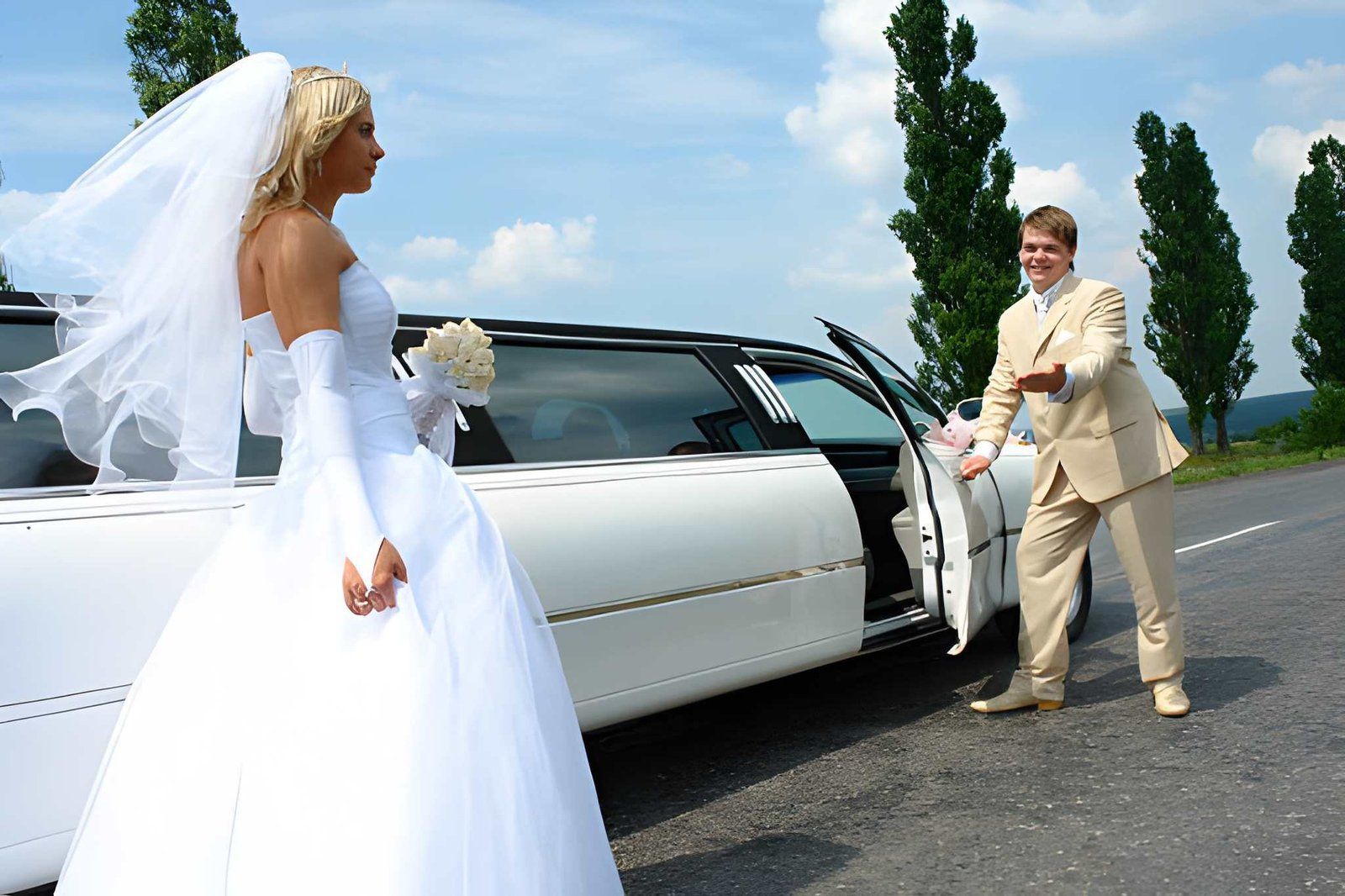 Bride in white gown by a white limousine, meeting a groom in a beige suit on a country road.