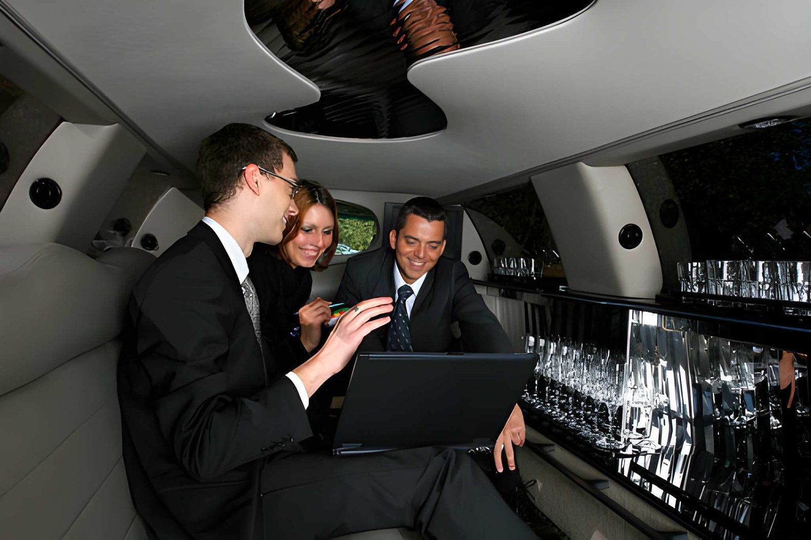 Three people in suits use a laptop inside a luxury limousine at night.