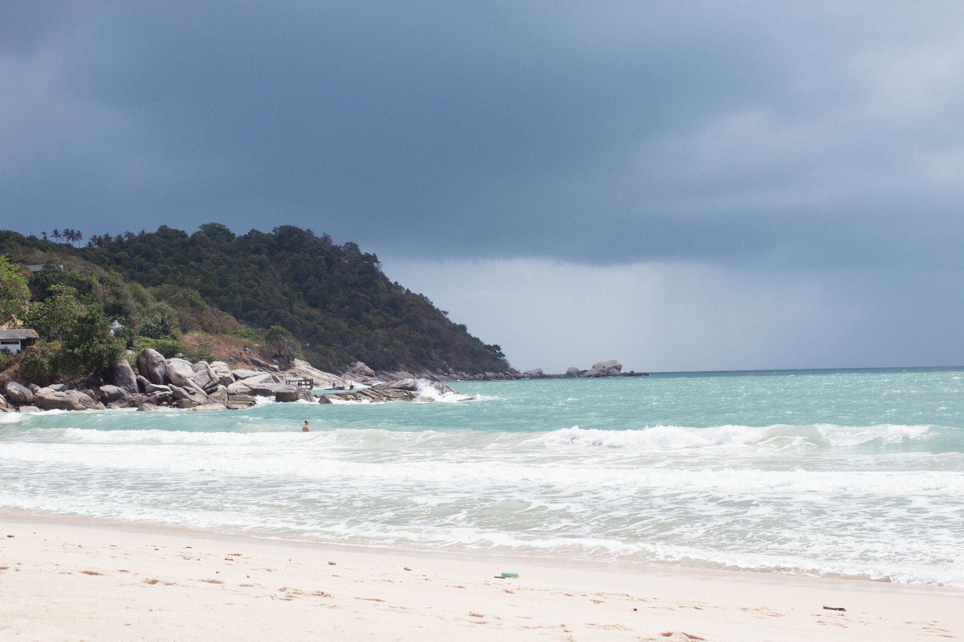 Stormy beach with turquoise waves, white sand, and a rocky, tree-covered headland under dark clouds