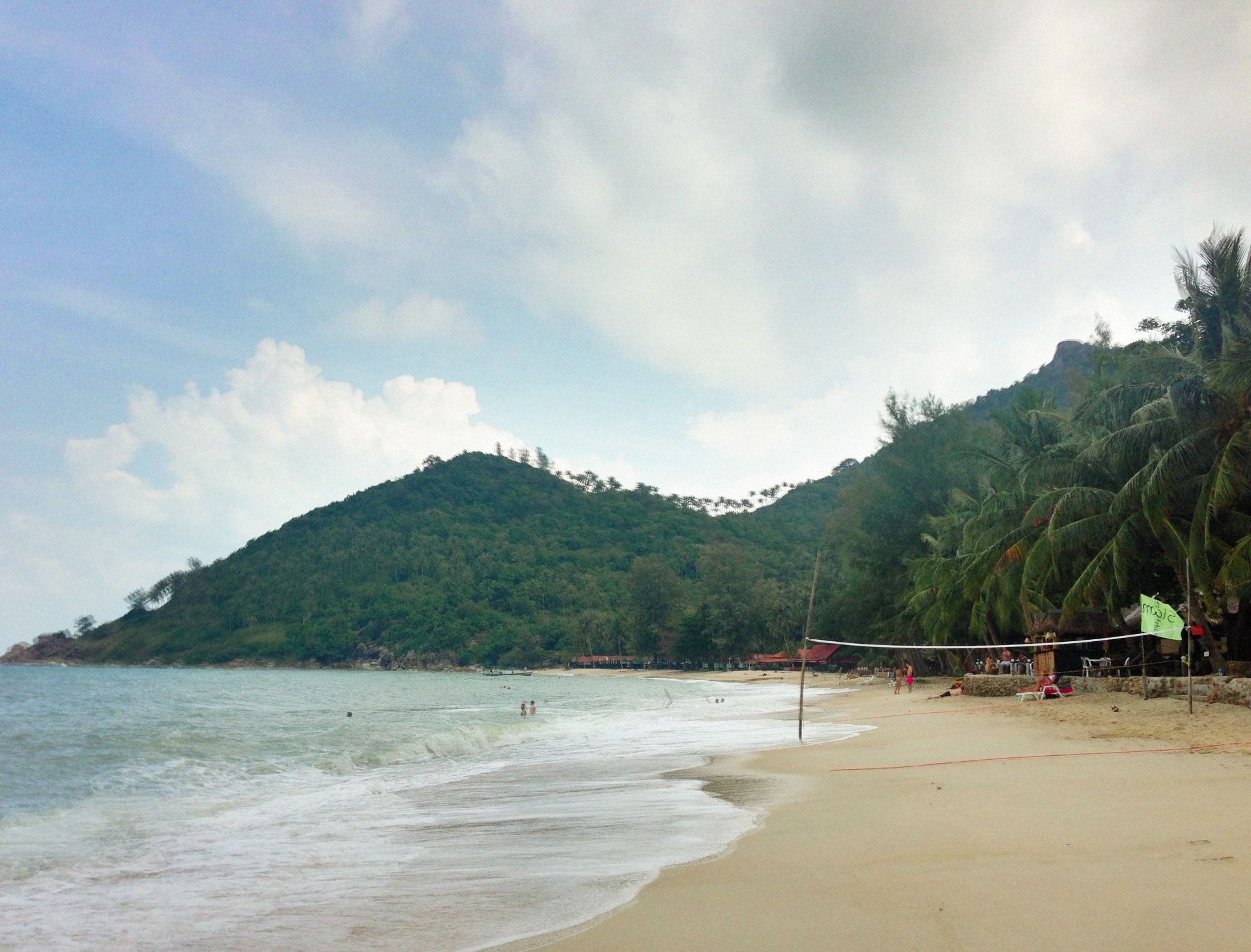 Sandy tropical beach with gentle waves, palm trees, and green hills under a cloudy sky